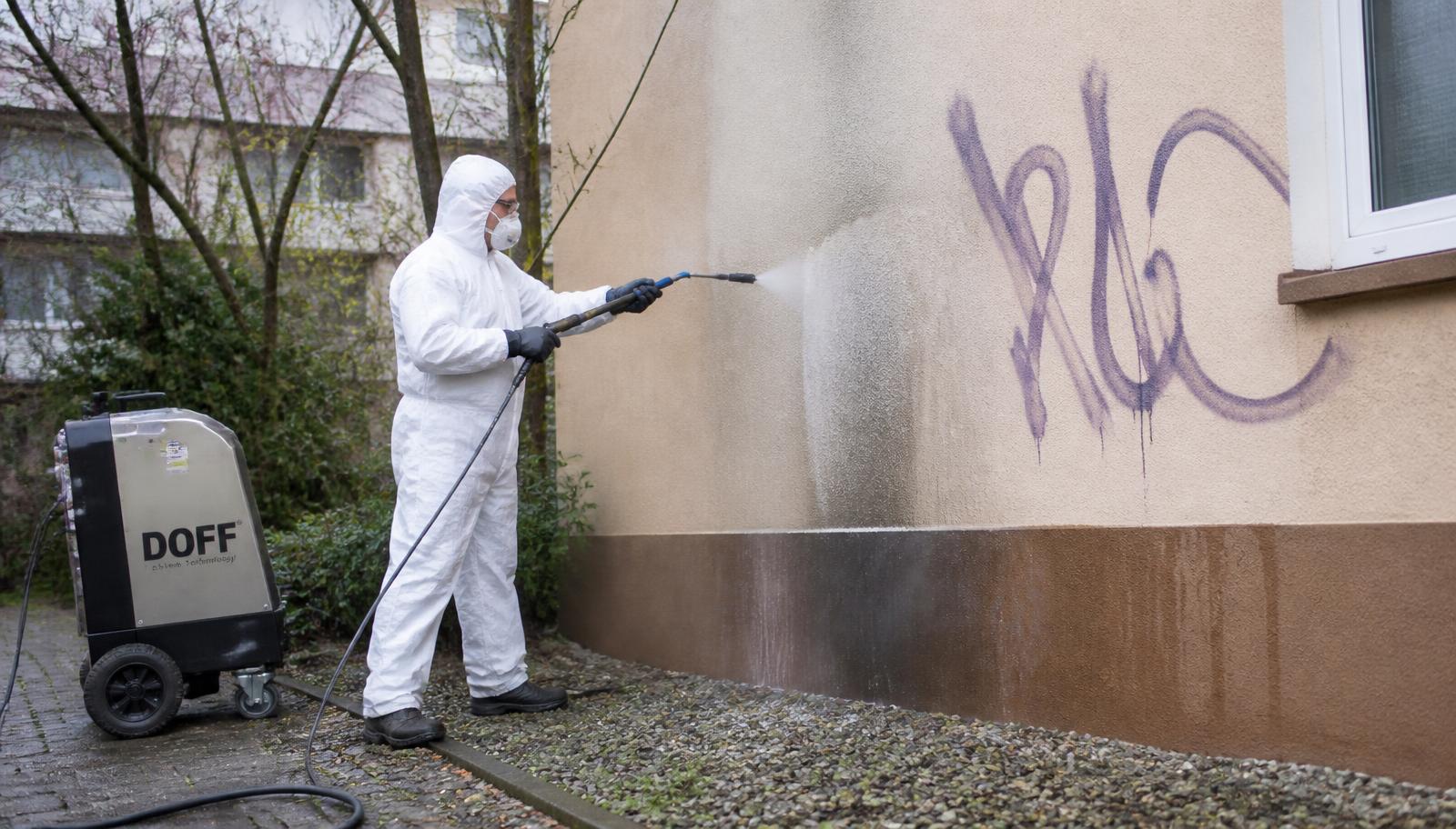 Fachkraft in Schutzanzug entfernt Graffiti mit einem Niederdruck-Heißwassergerät von einer Putzfassade